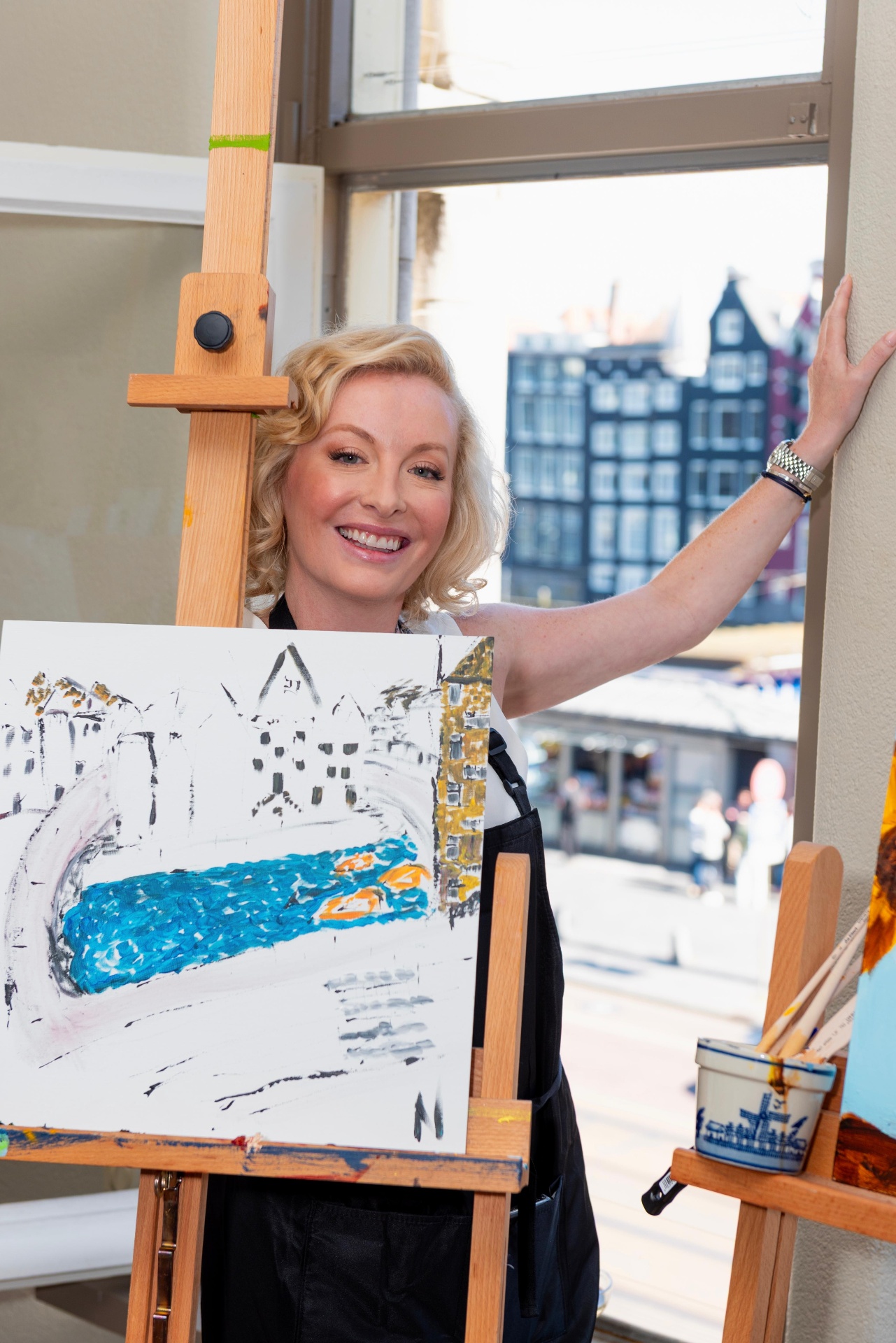 Smiling artist beside an easel with a fresh Amsterdam canal painting at Dutch Masters Studio